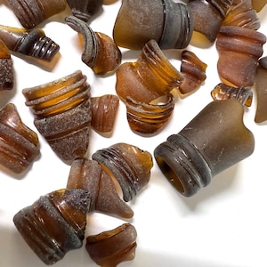 Könnte beinhalten: Eine Sammlung von bernsteinfarbenen, vom Meer geschliffenen Glasflaschenfragmenten. Die Teile variieren in Größe und Form, wobei einige Gewindehälse und abgerundete Kanten aufweisen. Das Glas hat ein mattiertes Aussehen, und die Fragmente sind auf einer weißen Oberfläche verstreut.