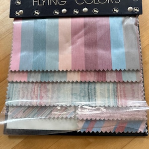 May include: A fabric swatch display featuring the text "FLYING COLORS" in white lettering. The display includes several fabric samples with vertical stripes in pastel shades of blue, pink, and lavender. The samples are attached to a black backing.