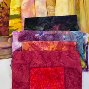 May include: A collection of colorful fabric pieces in various shades of red, purple, yellow, and brown. The fabrics appear to be dyed or painted with unique patterns and textures, suitable for crafting or textile projects.