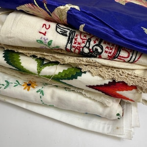 May include: A stack of embroidered textiles in various colors. A deep blue satin fabric with floral embroidery is on top. Below are white linens with colorful floral and geometric embroidery, and lace trim.