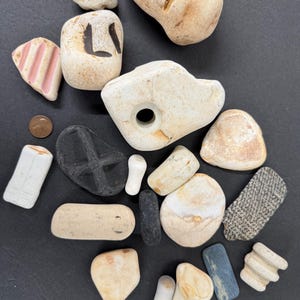 May include: A collection of small, weathered stones and ceramic fragments in beige, grey, black, and pink. Some pieces have markings or patterns, including a letter "L" and a cross. A penny is also included.