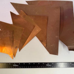 May include: Assortment of copper sheets in various shapes and sizes, arranged on a white surface. The sheets display a warm, reddish-brown hue. A black ruler with white markings is at the bottom, providing scale. The copper pieces have a metallic sheen.
