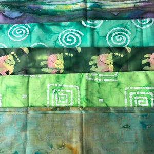 May include: A selection of batik fabric pieces in a stack. The fabrics display diverse patterns, such as swirls, geometric shapes, and organic designs. The colour scheme includes shades of green, blue, and brown, with white accents. The fabrics appear to be cotton or a similar material.