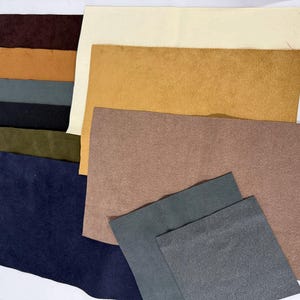 May include: A collection of various colored suede leather pieces. The colors include shades of brown, tan, olive green, navy blue, black, and gray. The leather pieces are rectangular and of varying sizes, suitable for crafting projects.