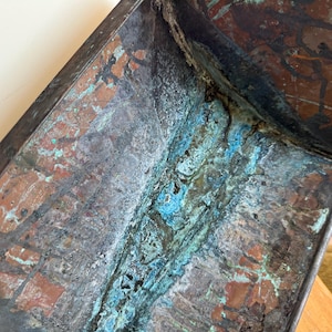 May include: A rectangular metal object with a weathered, oxidized surface featuring a mix of dark brown, copper, and vibrant turquoise green patina. The texture is rough and uneven, showing signs of age and corrosion across the metallic panels.