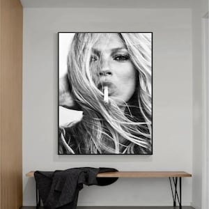 May include: Black and white photograph of a woman with long blonde hair, looking directly at the camera. She is holding a cigarette in her mouth.