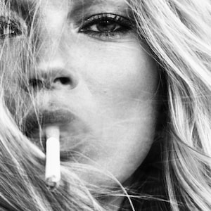 May include: A black and white portrait of a woman with long, flowing hair. She is looking off to the side and has a cigarette in her mouth.