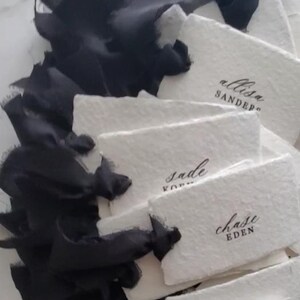 May include: A collection of white rectangular place cards with black handwritten names, tied with black silk ribbon. The names include "allisa sander", "sade", "chase eden", and "mackenzie babson". The cards have a textured, handmade appearance.