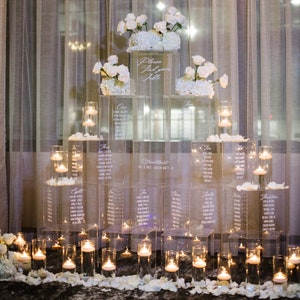 Puede incluir: Un plano de asientos de boda de acrílico transparente con arreglos florales blancos y velas flotantes. El gráfico muestra los nombres de los invitados y los números de mesa, con el texto "Please Find your Table". Un detalle elegante para la recepción.