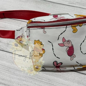 May include: White fanny pack with a red strap. The fanny pack has a Winnie the Pooh and Piglet print with yellow, red, and pink accents.