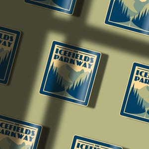 May include: Blue and white sticker with the text "The Icefields Parkway" in a vintage style font. The sticker features a mountain landscape with trees and a road.