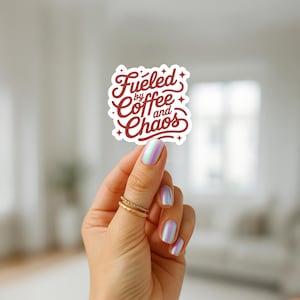 May include: A white sticker with the text "Fueled by Coffee and Chaos" in a burgundy script font. The sticker is held by a hand with iridescent nail polish and gold rings.