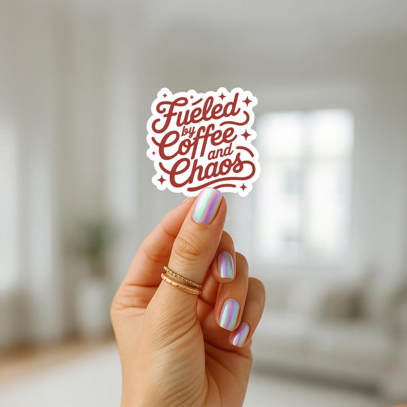 May include: A white sticker with the text "Fueled by Coffee and Chaos" in a burgundy script font. The sticker is held by a hand with iridescent nail polish and gold rings.