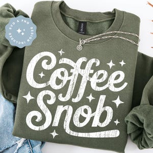 Puede incluir: Una sudadera verde oliva con el texto "Coffee Snob" en letras blancas desgastadas y acentos de estrellas. Un collar plateado con un colgante de sol está drapeado sobre la sudadera. Una pegatina azul dice "SVG & PNG DESIGN INCLUDED".