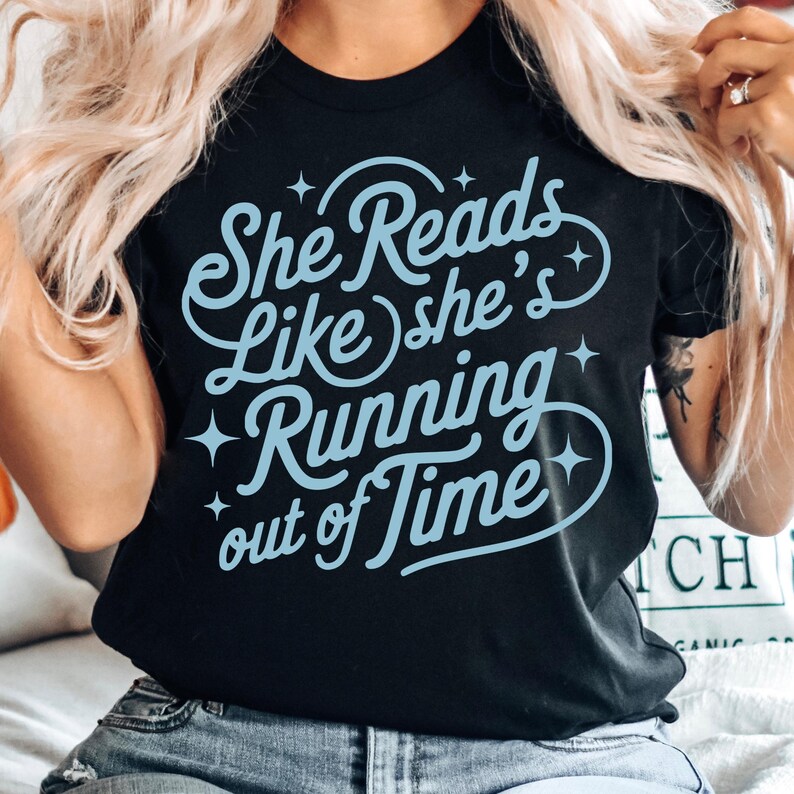 May include: Black t-shirt with the light blue text "She Reads Like she's Running out of Time". The text is in a whimsical font with star accents. The shirt is a crew neck.