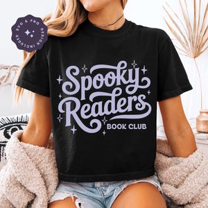 May include: A black short-sleeve t-shirt featuring the light purple text Spooky Readers BOOK CLUB in a stylized script font, accented by small four-pointed star graphics. A circular badge in the top left corner displays SVG & PNG DESIGN INCLUDED.