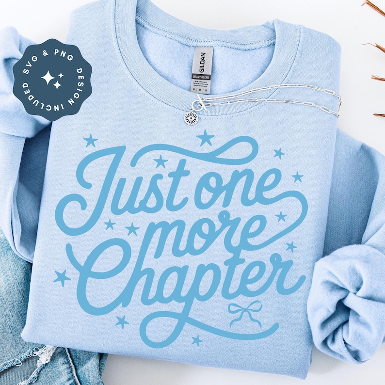 K&ouml;nnte beinhalten: Hellblaues Sweatshirt mit dem Text "Just one more chapter" in hellblauer Schrift. Das Design umfasst Sterne und eine Schleife. Eine silberne Halskette mit einem Blumenanh&auml;nger liegt auf dem Sweatshirt. Das Bild enth&auml;lt auch eine Grafik "SVG & PNG DESIGN INCLUDED".