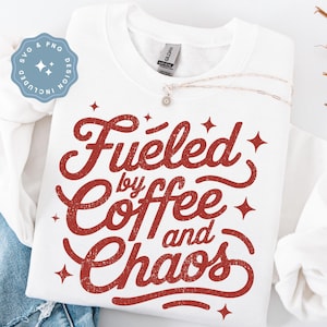 May include: White sweatshirt with the phrase "Fueled by Coffee and Chaos" in red, distressed lettering. The design includes star accents. A silver necklace with a small pendant is draped across the sweatshirt. The image also includes a blue and white SVG and PNG design.