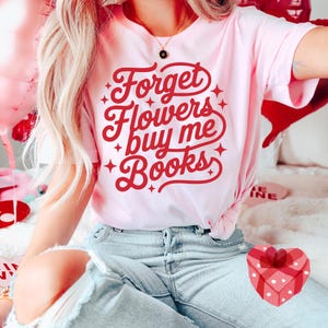Puede incluir: Camiseta rosa claro con el texto rojo "Forget Flowers buy me Books". La camiseta se usa con vaqueros azul claro. Una caja de regalo roja en forma de coraz&oacute;n es visible.