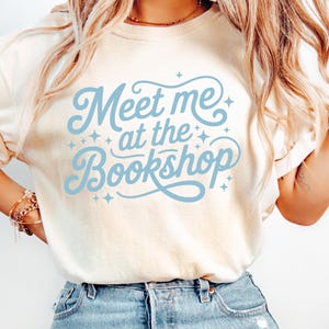 May include: Cream-colored t-shirt with the text "Meet me at the Bookshop" in light blue script. The design includes decorative star accents. The shirt is a casual style, suitable for everyday wear.