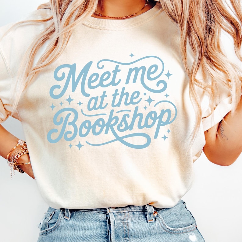 May include: Cream-colored t-shirt with the text "Meet me at the Bookshop" in light blue script. The design includes decorative star accents. The shirt is a casual style, suitable for everyday wear.