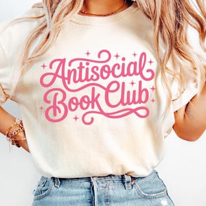 K&ouml;nnte beinhalten: Cremefarbenes T-Shirt mit dem pinkfarbenen Schriftzug "Antisocial Book Club" in verschn&ouml;rkelter Schrift. Der Text ist von kleinen Sternen umgeben. Das Shirt besteht aus weichem Material und hat kurze &Auml;rmel.