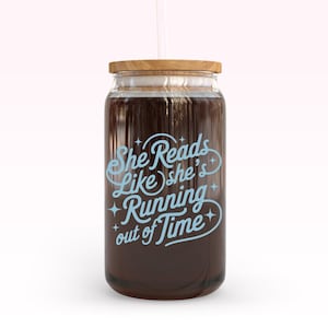 May include: A clear glass jar with a wooden lid and a straw. The jar is filled with a dark liquid and features the text "She Reads Like she's Running out of Time" in light blue script.