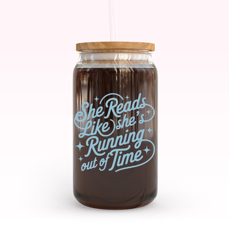 May include: A clear glass jar with a wooden lid and a straw. The jar is filled with a dark liquid and features the text "She Reads Like she's Running out of Time" in light blue script.