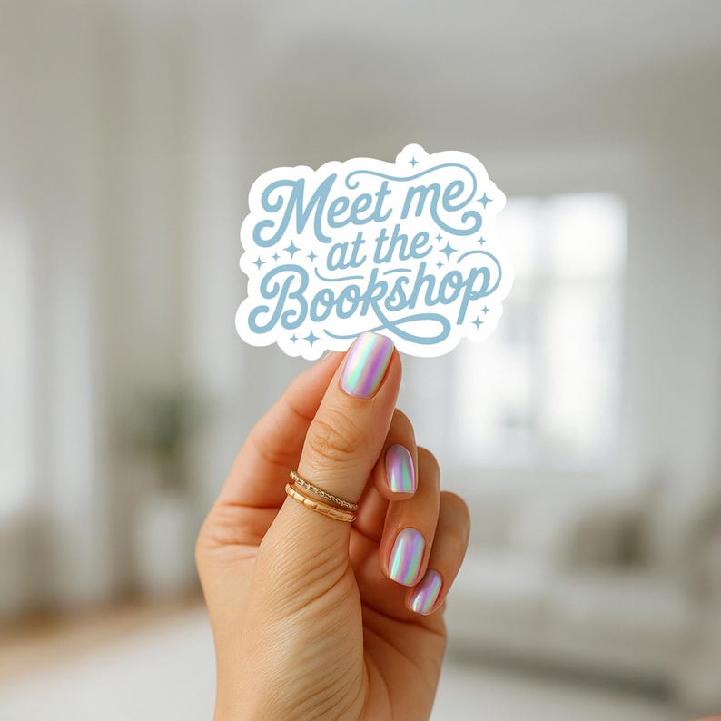 May include: A white sticker with the text "Meet me at the Bookshop" in light blue script. The sticker is held up by a hand with iridescent nail polish and gold rings.