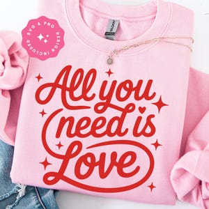 Puede incluir: Sudadera rosa claro con la frase "All you need is Love" en rojo. El diseño incluye estrellas y corazones. Un collar dorado con un colgante está sobre la sudadera.