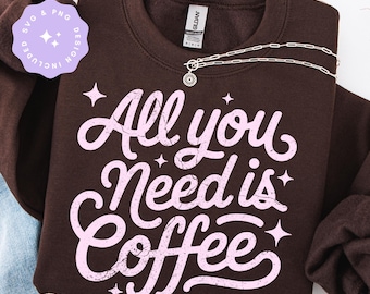 May include: A brown sweatshirt with the phrase "All you Need is Coffee" in light pink script. A silver necklace with a sun pendant is draped across the front. A small, round sticker with text is in the upper left corner.