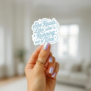 May include: A white sticker with the text "She Reads Like she's Running out of Time" in a light blue script. The sticker is held up by a hand with iridescent nail polish and gold rings.