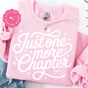 K&ouml;nnte beinhalten: Ein hellrosa Sweatshirt mit dem wei&szlig;en Schriftzug "Just one more chapter". Das Design beinhaltet Sterne und eine Schleife. Eine goldene Halskette mit einem Blumenanh&auml;nger liegt auf dem Sweatshirt. Eine rosa SVG & PNG Design-Grafik befindet sich in der oberen linken Ecke.