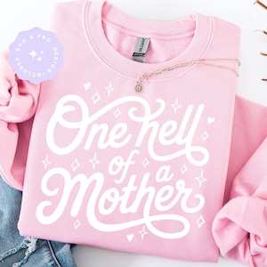 May include: A light pink sweatshirt with the phrase "One Hell of a Mother" in white script. The design includes stars and hearts. A gold necklace with a flower pendant is draped across the sweatshirt. The words "SVG & PNG DESIGN INCLUDED" are in a purple circle.