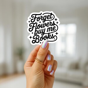 Puede incluir: Una pegatina blanca con el texto "Forget Flowers buy me Books" en una fuente negra y estilizada. La pegatina est&aacute; sujeta por una mano con esmalte de u&ntilde;as iridiscente y anillos dorados.