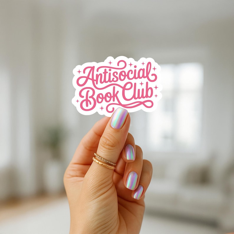 K&ouml;nnte beinhalten: Ein wei&szlig;er Aufkleber mit dem Text "Antisocial Book Club" in rosa Schrift. Der Aufkleber wird von einer Hand mit schillerndem Nagellack und goldenen Ringen gehalten. Das Design weist dekorative Verzierungen und Sterne auf.
