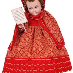 May include: A detailed image of a doll dressed in a red and gold patterned dress and cape. The doll holds a scroll with text. The doll has brown hair and green eyes. The dress has a scalloped hemline.