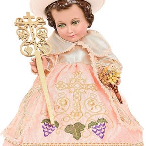 May include: A detailed doll of a child figure, wearing a pink dress with gold embroidery and lace trim. The doll holds a gold cross-shaped scepter and a bouquet of flowers. It has a white hat and a cream-colored cape.