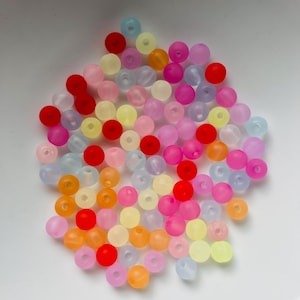 Könnte beinhalten: Eine Ansammlung kleiner, runder Perlen mit matter Oberfläche in verschiedenen Farben wie Rot, Orange, Gelb, Hellblau, Pink und Lila. Die Perlen sind in einer Gruppe verstreut und weisen jeweils ein kleines Loch in der Mitte zum Auffädeln auf.