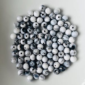 Könnte beinhalten: Eine Ansammlung runder Perlen mit einem Marmormuster in Weiß-, Grau- und Schwarztönen. Jede Perle hat ein mittiges Loch und die Oberfläche der Perlen weist ein glattes, glänzendes Finish mit wirbelnden Farbvariationen im gesamten Set auf.