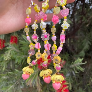 May include: A collection of beaded phone charms in shades of yellow, pink, and clear. The charms feature various shapes, including flowers, hearts, and paw prints, with small yellow dog figures holding pink hearts at the bottom. The charms are strung on pink cords.