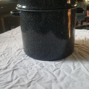 May include: A black enamel stockpot with a speckled white finish, featuring two side handles, a matching lid with a central handle, and a secondary insert section. The cylindrical pot has a glossy surface and a sturdy construction for cooking.
