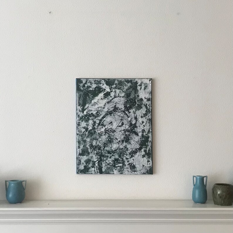 May include: Abstract painting in shades of black and white with a textured surface. The painting is framed and hanging on a white wall above a mantelpiece.