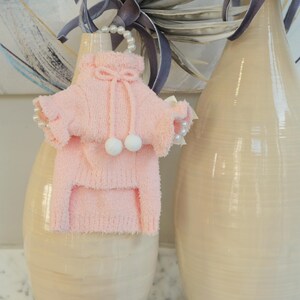 May include: A light pink pet sweater with a hood and white pom-poms. The sweater has a drawstring and is displayed on a beige vase. A pearl necklace is draped over the vase and sweater. The sweater is made of a soft, fuzzy material.