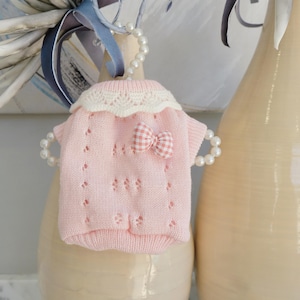 May include: A small, pink knitted sweater with a white lace collar and a gingham bow. The sweater has a delicate openwork design and is displayed on a pearl beaded hanger. The overall theme is baby clothing.
