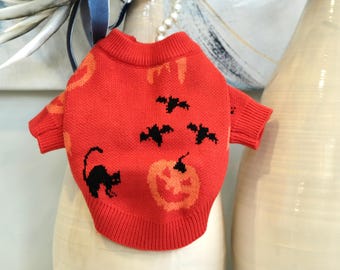 May include: A red Halloween-themed dog sweater with black cat and bat designs, and a jack-o'-lantern. The sweater is made of a knitted material.