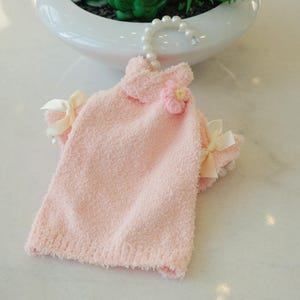 May include: A light pink pet dress with a textured, fluffy fabric. The dress features a small flower accent and cream-colored bows on the sleeves. It hangs from a pearl-beaded hanger.