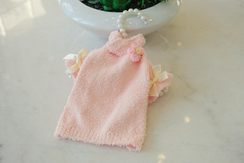 May include: A light pink pet dress with a textured, fluffy fabric. The dress features a small flower accent and cream-colored bows on the sleeves. It hangs from a pearl-beaded hanger.