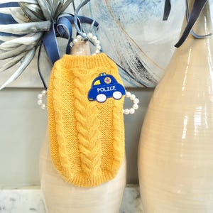 Puede incluir: Un suéter amarillo mostaza para perros tejido con trenzas, con un parche azul de coche de policía. El parche tiene la palabra "POLICE" y una estrella. El suéter se exhibe en un jarrón crema, con una cinta azul y detalles de perlas.