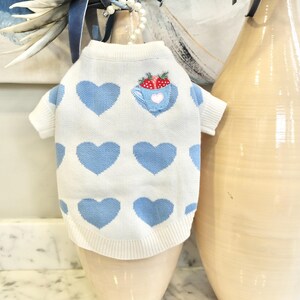 May include: A white dog sweater with light blue heart patterns. The sweater has short sleeves and a ribbed hem. A small patch on the chest features a teacup with strawberries and a heart.